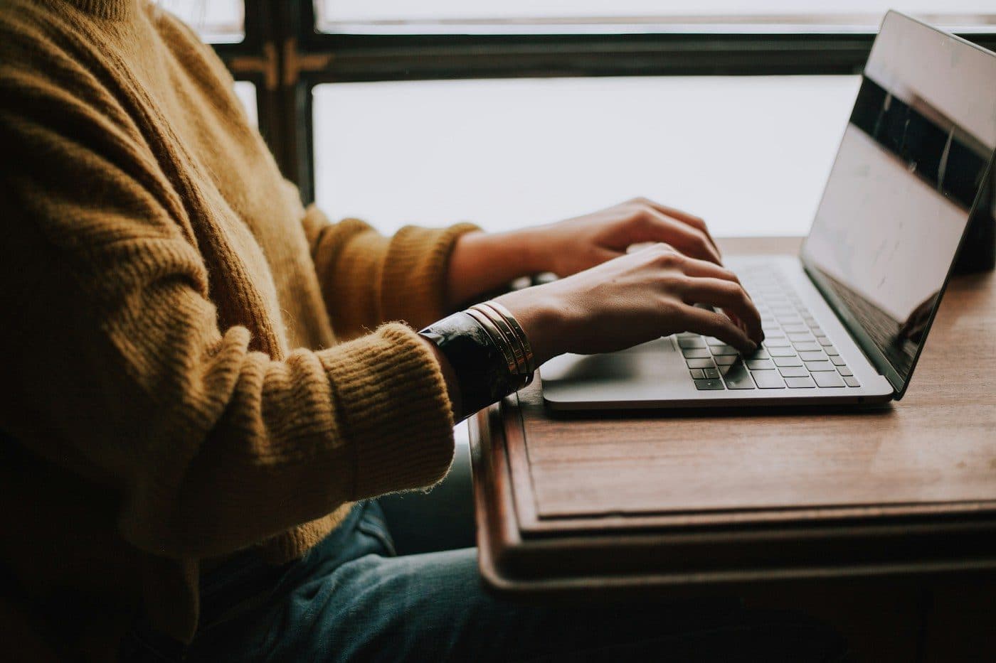 A founder in a mustard jumper typing on a laptop at a wooden desk by a window