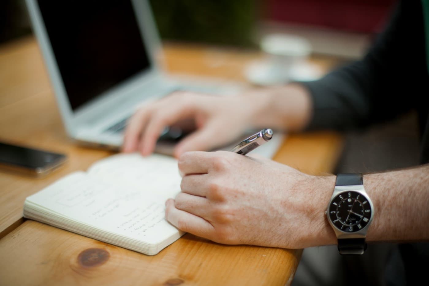 A hand writing in a notebook with a pen beside a laptop on a wooden table